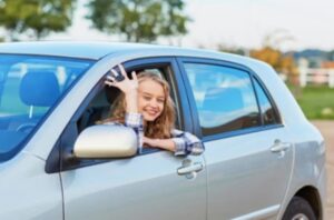 A smiling young woman waving from the driver's seat of a car, assisted by a 24-hour locksmith to regain access.
