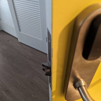 Close-up of a yellow door slightly ajar with a stainless steel handle, showing a wooden floor and a white louvered door in the background. A mobile locksmith is on the way to rekey