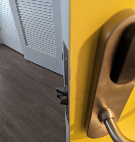 Close-up of a yellow door slightly ajar with a stainless steel handle, showing a wooden floor and a white louvered door in the background. A mobile locksmith is on the way to rekey